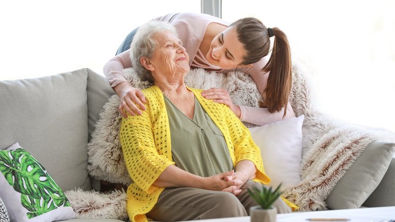 Eine Enkelin mit rosanem Oberteil und braunem langen Haar schaut von hinten auf ihre Großmutter die auf einem hellen Sofa sitzt. Die Großmutter trägt eine olivfarbene Bluse und gelbe Weste.