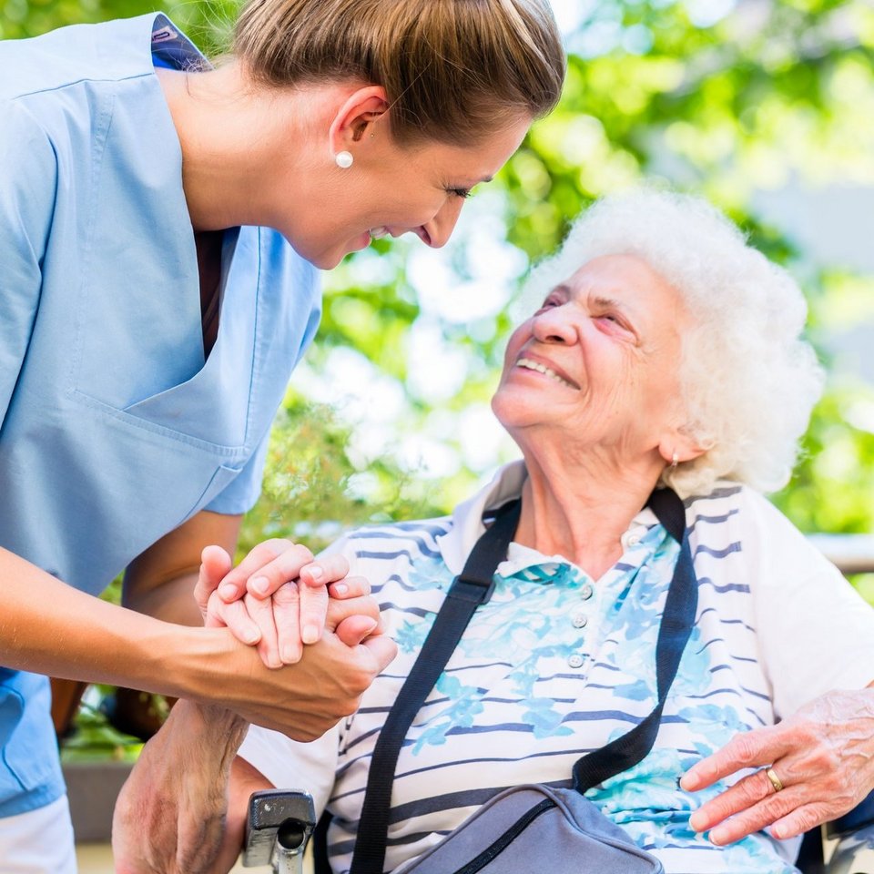 Eine Pflegerin mit braunem Haar hält die Hand einer Bewohnerin des Pflegeheims. Die Bewohnerin sitzt im Rollstuhl draußen auf einer Terrasse.