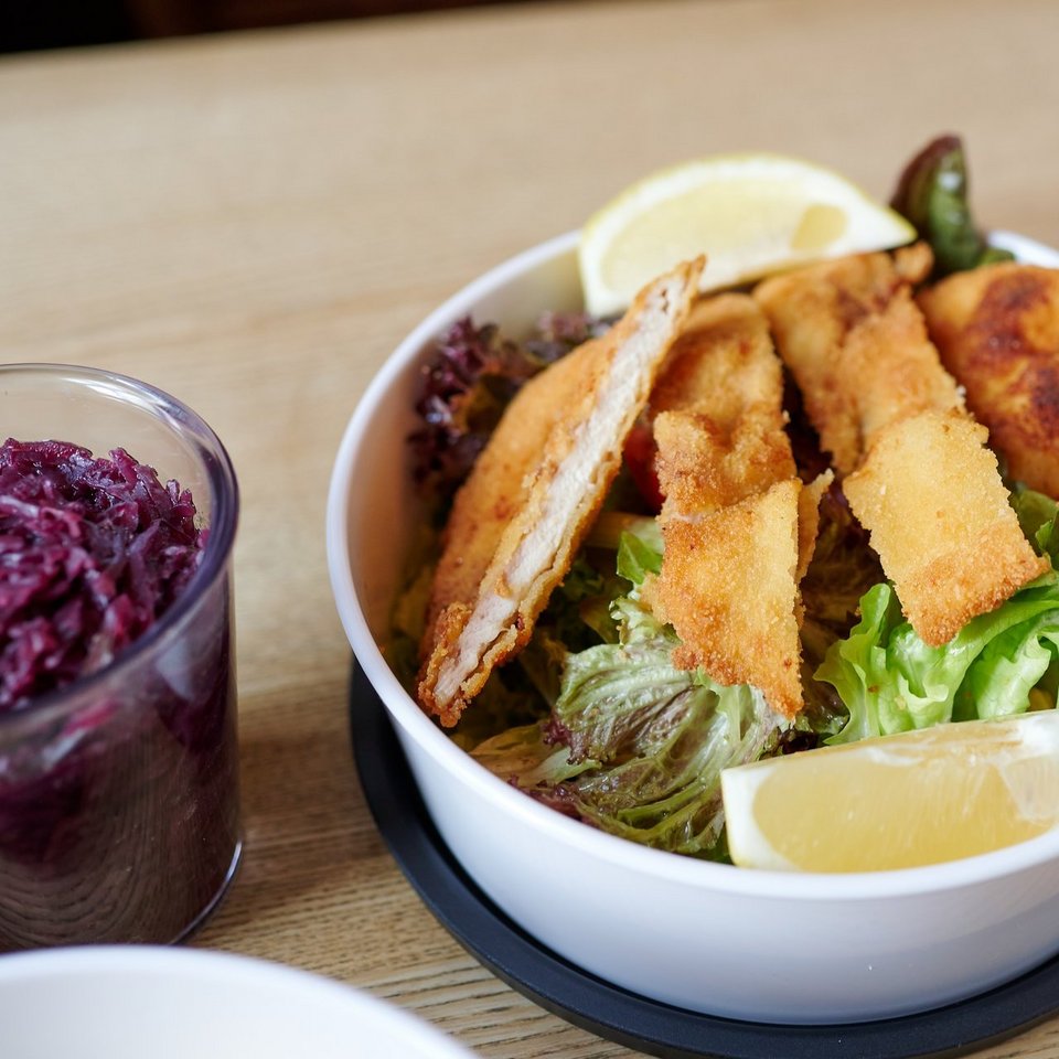 Ein bunter Salat mit einem geschnittenen Schnitzel steht in einer To-Go-Schüssel auf einem Holztisch. Links neben dem Salat steht ein Glas mit Rotkohl.