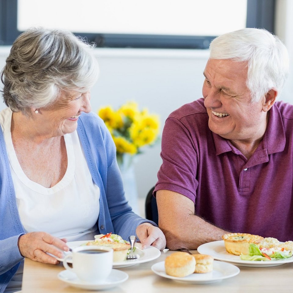 Zwei Senioren sitzen an einem gedeckten Tisch und lächeln sich an.