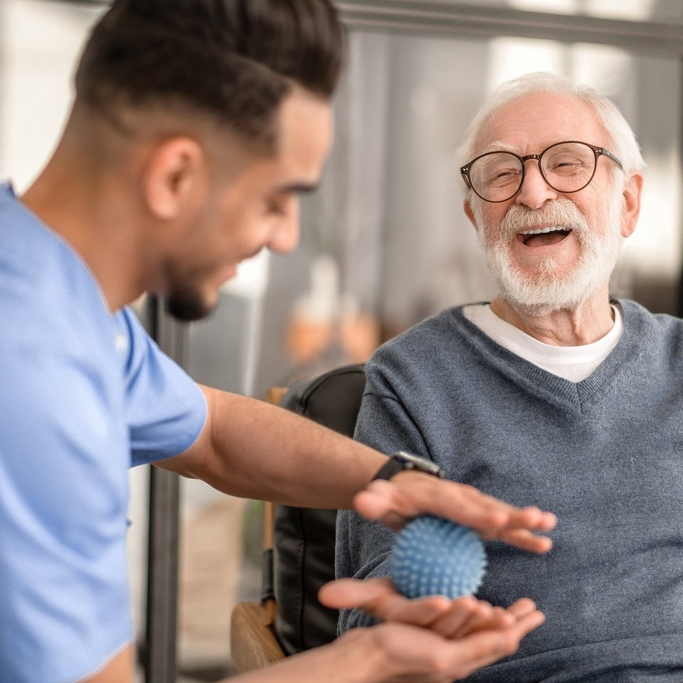 Ein fröhlicher alter Mann unterzieht sich einer Physiotherapie-Sitzung der Physiotherapie, die von einem Pfleger durchgeführt wird.