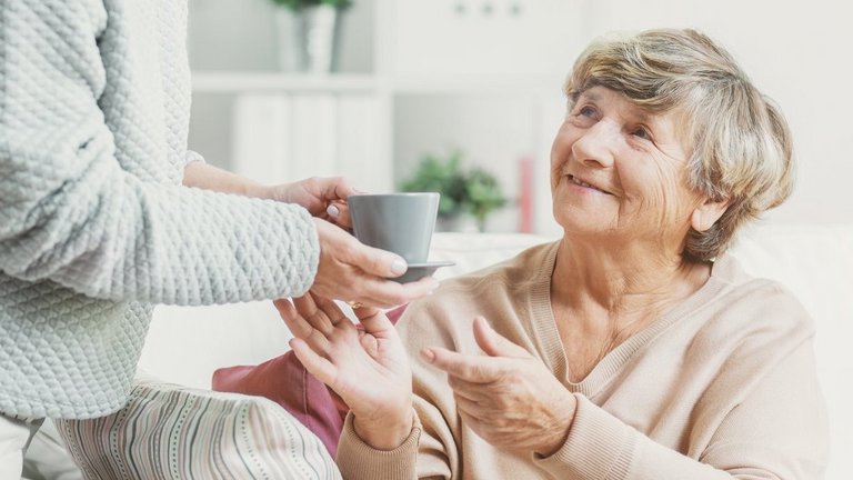 Nahaufnahme einer Person die einer Seniorin eine Teetasse reicht. Die Seniorin sitzt auf einem Sofa und schaut die andere Person an.