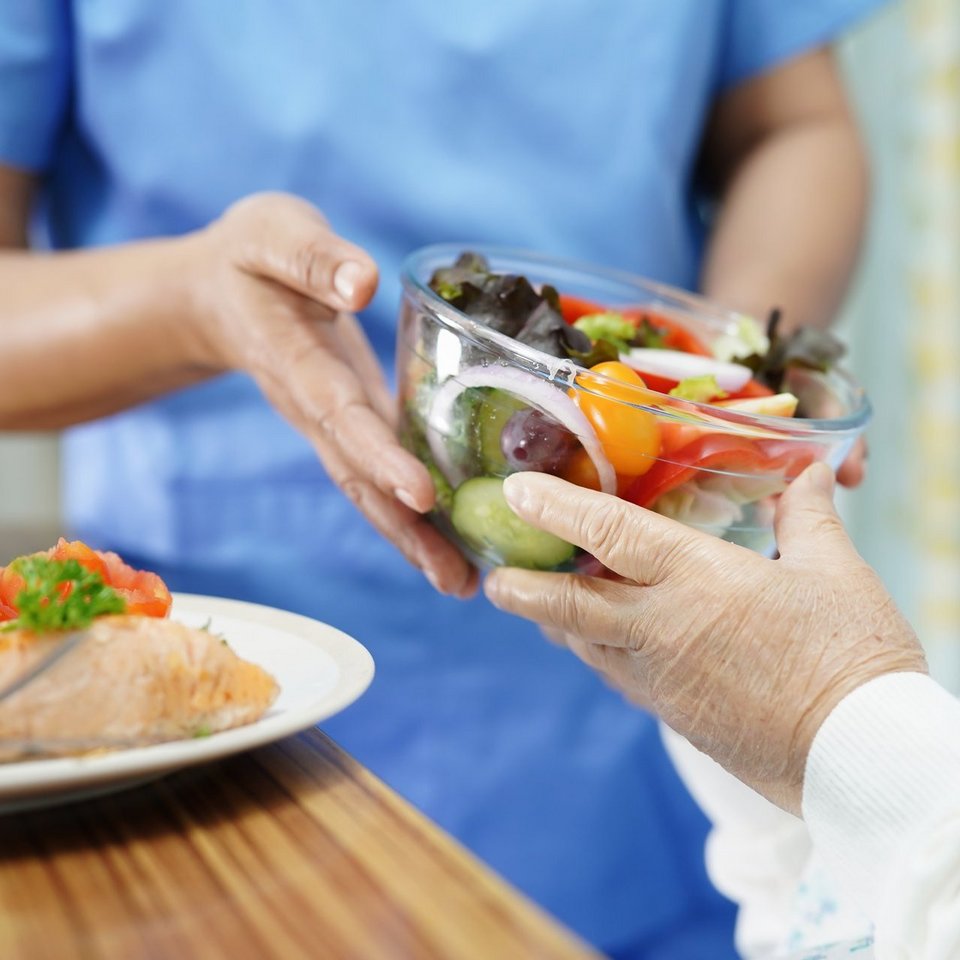 Eine Pflegerin reicht einer Senioren eine Schüssel mit frischem Salat. Auf dem Tisch vor den beiden steht ein Teller mit Fleisch und gekochtem Gemüse.