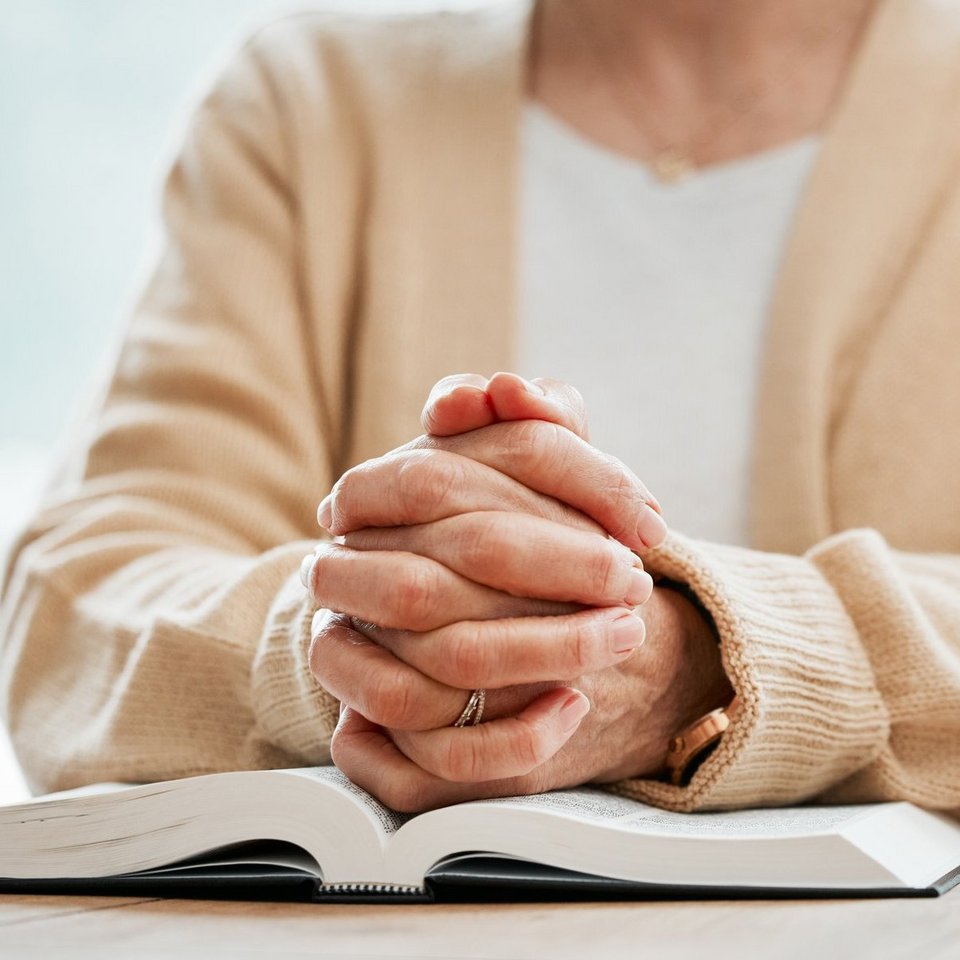 Eine Seniorin gekleidet mit einem hellen Oberteil legt ihre gefalteten Hände auf eine aufgeschlagene Bibel.