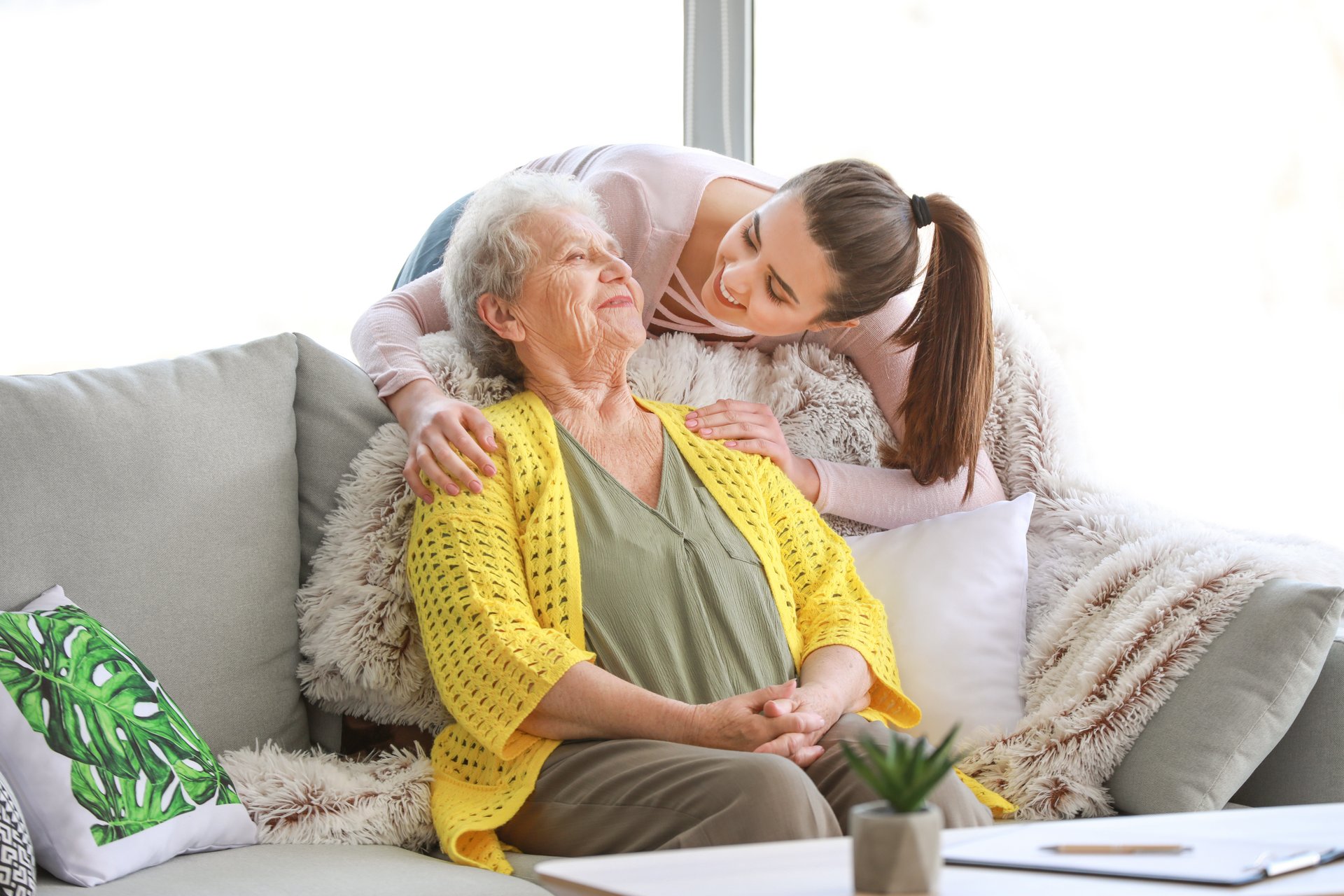 Eine Enkelin mit rosanem Oberteil und braunem langen Haar schaut von hinten auf ihre Großmutter die auf einem hellen Sofa sitzt. Die Großmutter trägt eine olivfarbene Bluse und gelbe Weste.