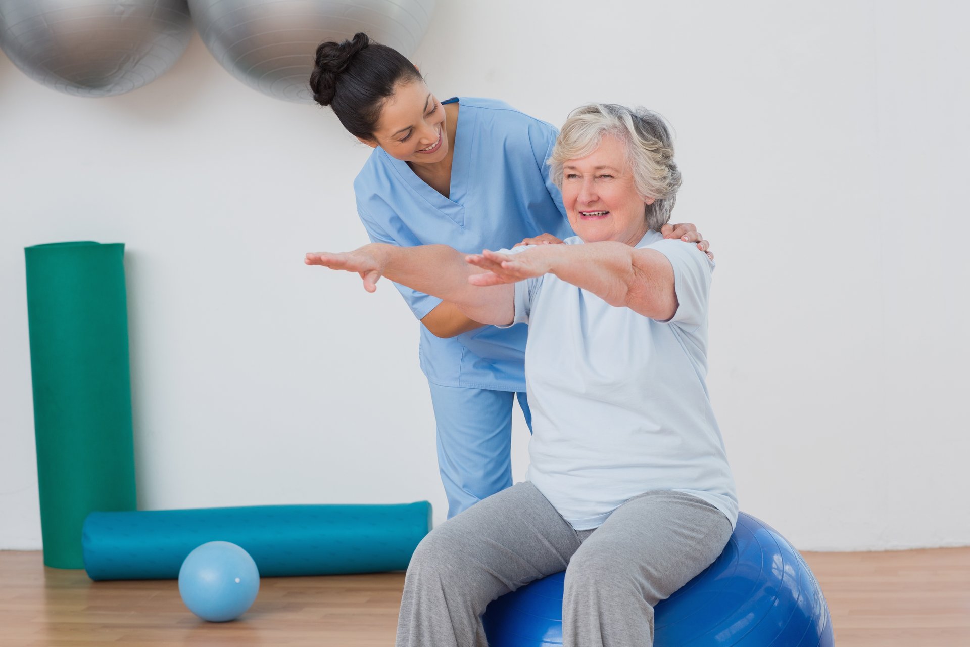 Eine Pflegerin instruiert eine ältere Frau, die auf im Sport-Outfit auf einem blauen Gymnastikball sitzt. Die beiden Frauen befinden sich in einer Sporthalle.